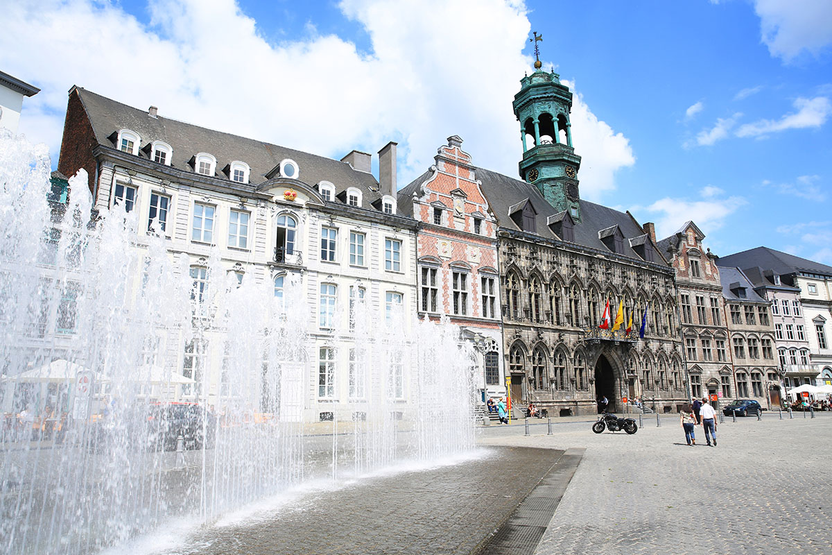A cobbled town square with Gothic buildings lining it and fountains spraying water